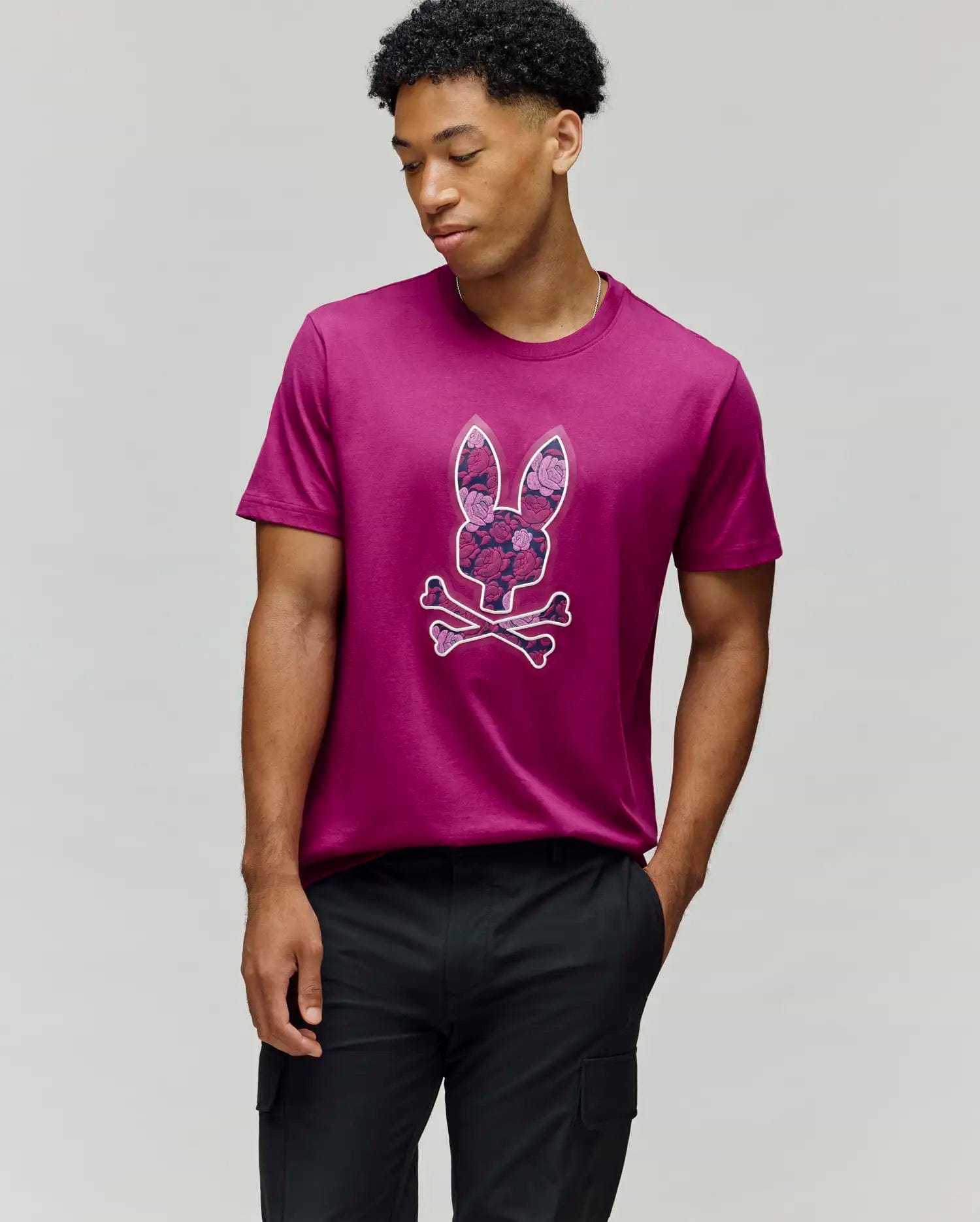 A man stands against a plain gray background wearing the Psycho Bunny MENS ROWEN BUNNY GRAPHIC TEE (MT0200101), a magenta Pima cotton shirt with a bunny skull and crossbones graphic, one hand in his pocket as he looks down to the side. (2)