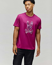 A young man wears the Psycho Bunny MENS ROWEN BUNNY GRAPHIC TEE - MT0200101, featuring a large bunny head above crossed bones, paired with black cargo pants against a plain light gray background. (4)
