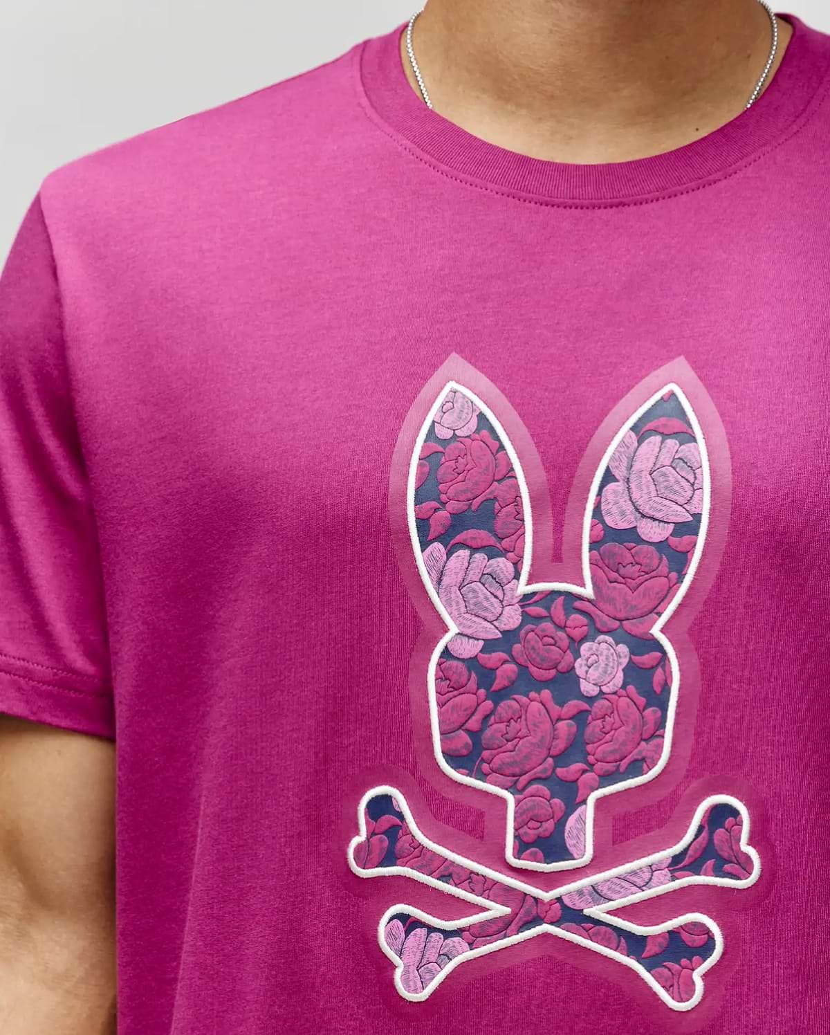 A person wearing the Psycho Bunny MENS ROWEN BUNNY GRAPHIC TEE (MT0200101), featuring a large bunny head and crossbones with a pink and purple floral pattern. Only the upper torso is visible. (3)