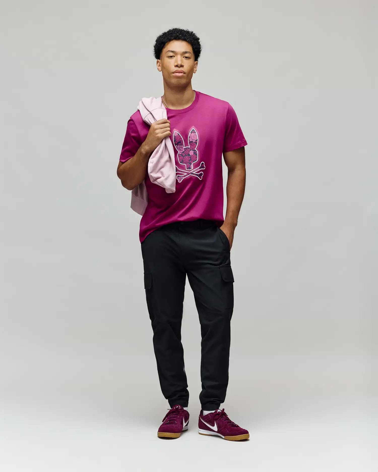 A man stands against a gray background wearing the Psycho Bunny MENS ROWEN BUNNY GRAPHIC TEE - MT0200101 in magenta, black cargo pants, and maroon sneakers. He holds a pink jacket over his shoulder and looks confidently at the camera. (5)