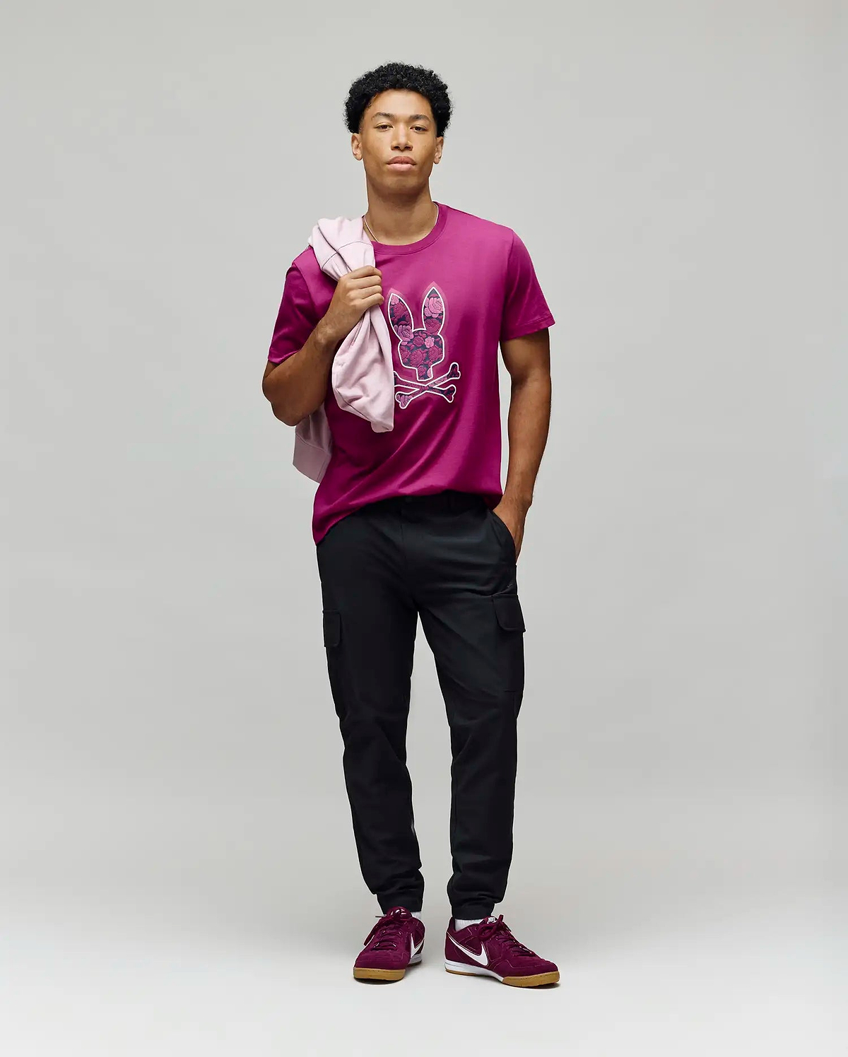 A man stands against a gray background wearing the Psycho Bunny MENS ROWEN BUNNY GRAPHIC TEE - MT0200101 in magenta, black cargo pants, and maroon sneakers. He holds a pink jacket over his shoulder and looks confidently at the camera. (5)