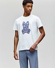 A young man wears the Psycho Bunny MENS ROWEN BUNNY GRAPHIC TEE (MT0200101), featuring a large bunny head and crossbones graphic on the front, paired with light blue pants, standing against a plain light gray background. (2)