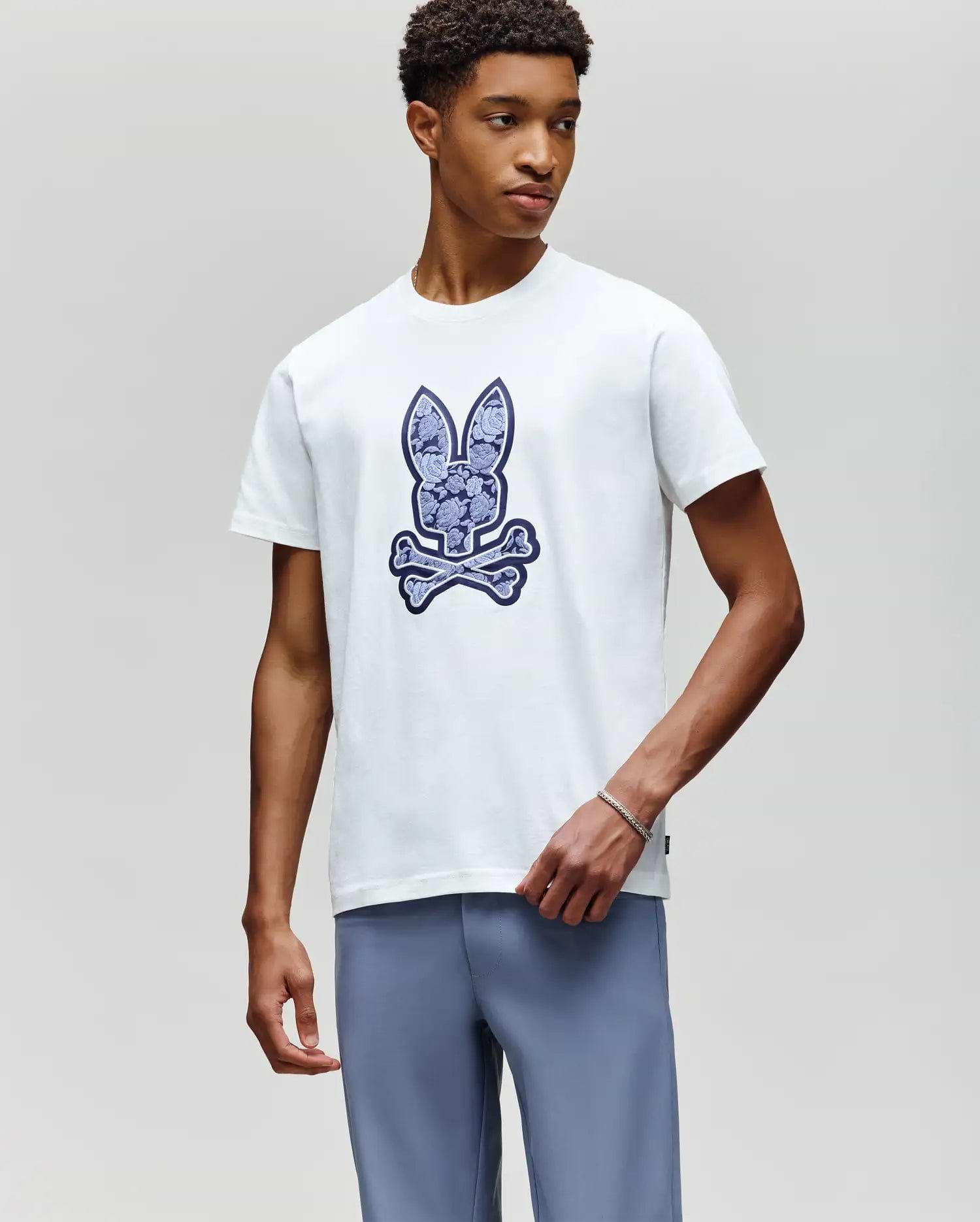A young man wears the Psycho Bunny MENS ROWEN BUNNY GRAPHIC TEE (MT0200101), featuring a large bunny head and crossbones graphic on the front, paired with light blue pants, standing against a plain light gray background. (2)