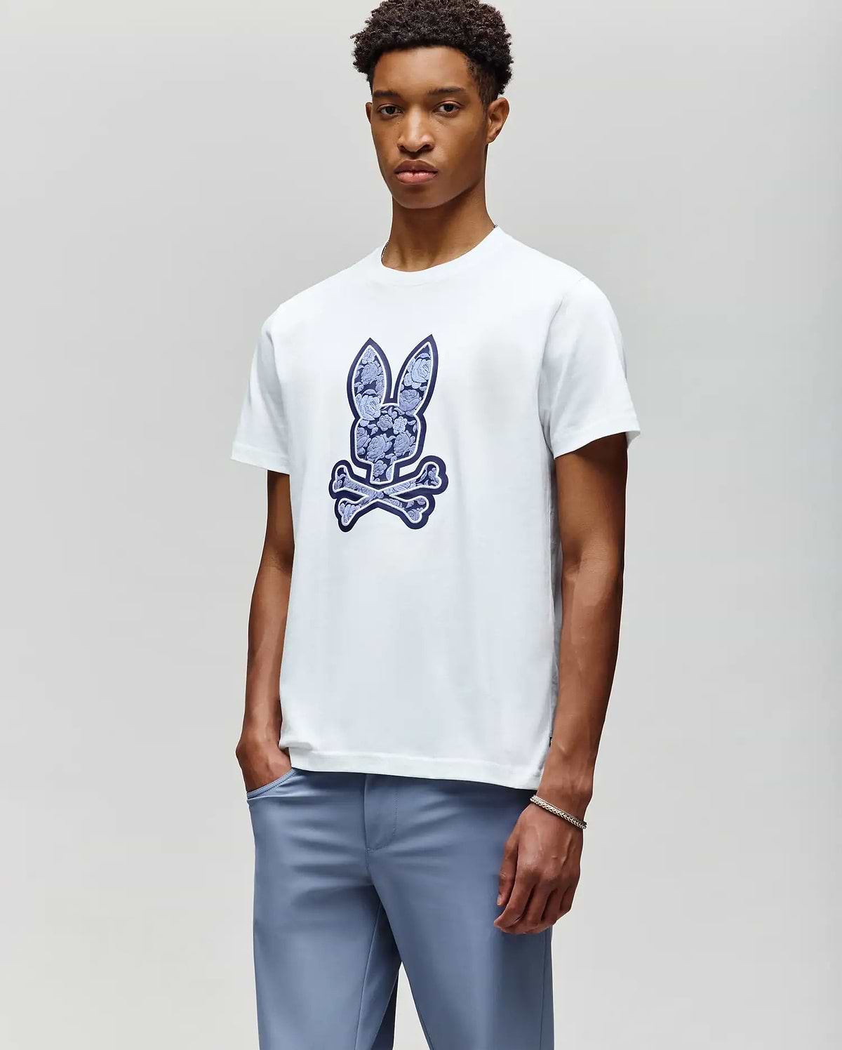 A young man wears the Psycho Bunny MENS ROWEN BUNNY GRAPHIC TEE (MT0200101), a white t-shirt featuring a large blue bunny head above crossed bones, paired with light blue pants, standing against a plain background. (4)