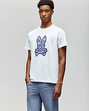 A young man wears the Psycho Bunny MENS ROWEN BUNNY GRAPHIC TEE (MT0200101), a white t-shirt featuring a large blue bunny head above crossed bones, paired with light blue pants, standing against a plain background. (4)