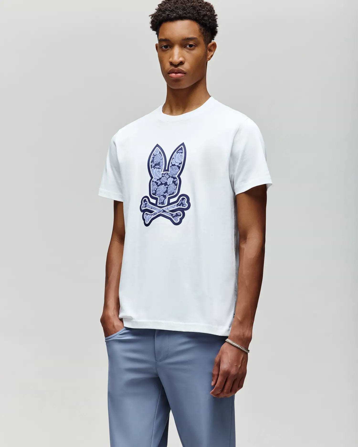 A young man wears the Psycho Bunny MENS ROWEN BUNNY GRAPHIC TEE (MT0200101), a white t-shirt featuring a large blue bunny head above crossed bones, paired with light blue pants, standing against a plain background. (4)