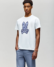 A young man wears the Psycho Bunny MENS ROWEN BUNNY GRAPHIC TEE (MT0200101), a white t-shirt featuring a large blue bunny head above crossed bones, paired with light blue pants, standing against a plain background. (4)
