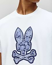 A person wears the Psycho Bunny MENS ROWEN BUNNY GRAPHIC TEE (MT0200101), a white t-shirt featuring a blue floral-patterned bunny skull and crossbones graphic. Only their lower face and neck are shown. (3)