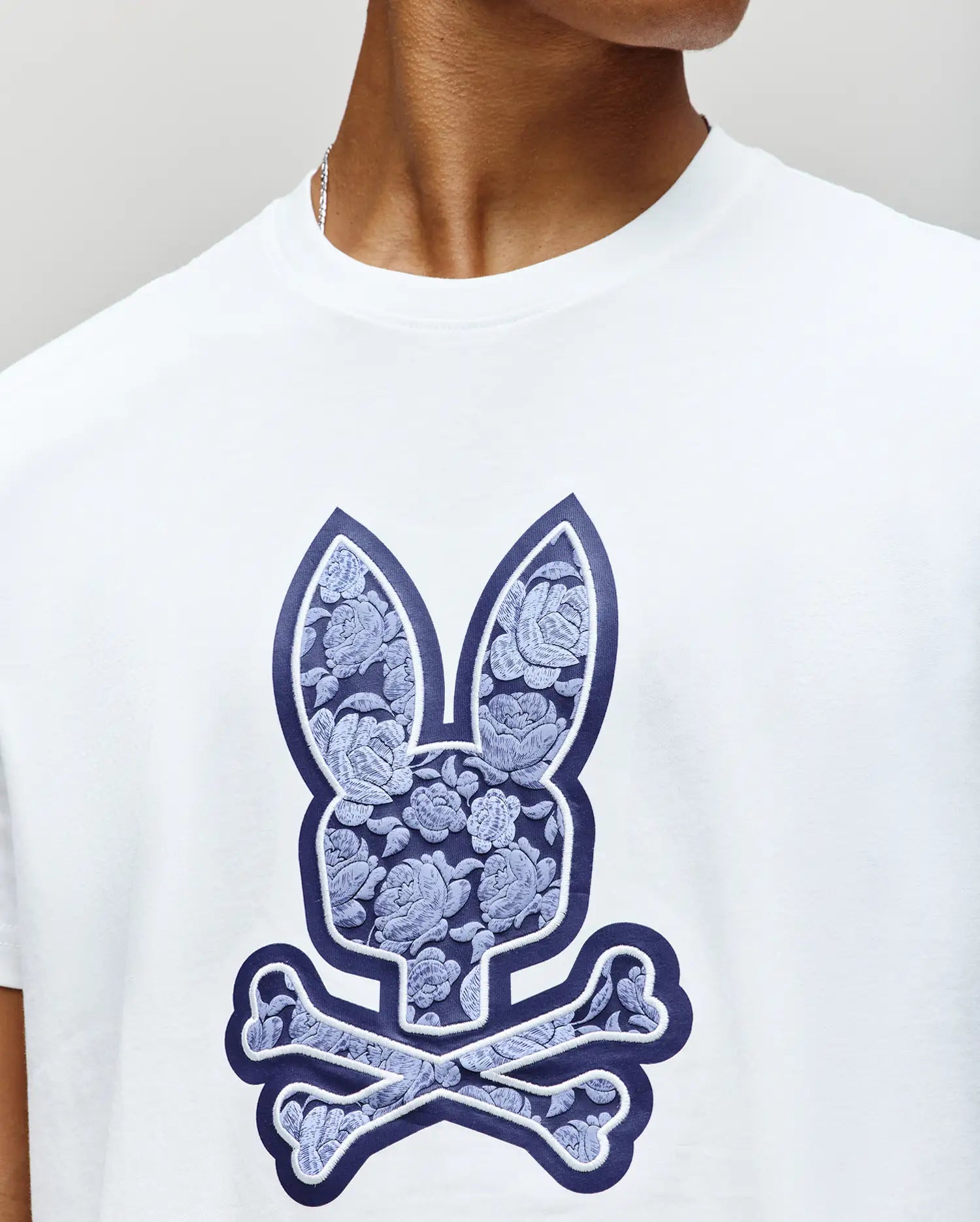A person wears the Psycho Bunny MENS ROWEN BUNNY GRAPHIC TEE (MT0200101), a white t-shirt featuring a blue floral-patterned bunny skull and crossbones graphic. Only their lower face and neck are shown. (3)