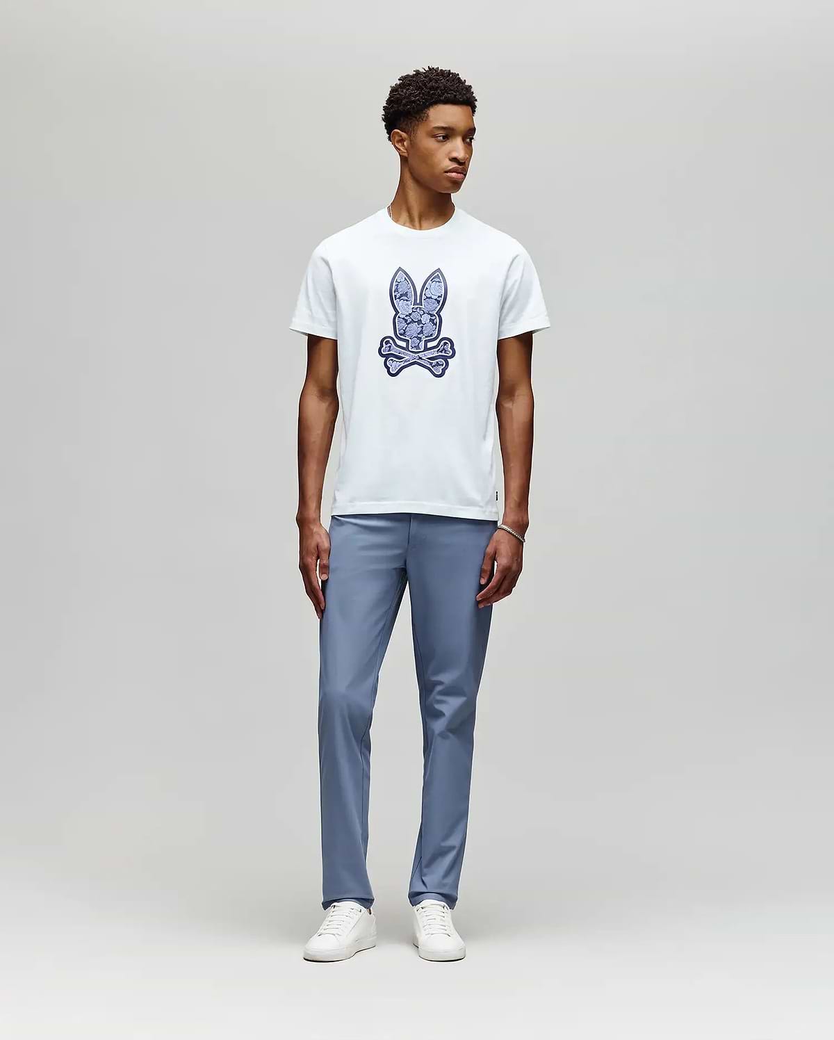 A young man stands against a light gray background, wearing the Psycho Bunny MENS ROWEN BUNNY GRAPHIC TEE (MT0200101) in white with a blue graphic, paired with light blue pants and white sneakers, looking slightly left with a neutral expression. (5)