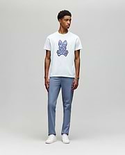 A young man stands against a light gray background, wearing the Psycho Bunny MENS ROWEN BUNNY GRAPHIC TEE (MT0200101) in white with a blue graphic, paired with light blue pants and white sneakers, looking slightly left with a neutral expression. (5)