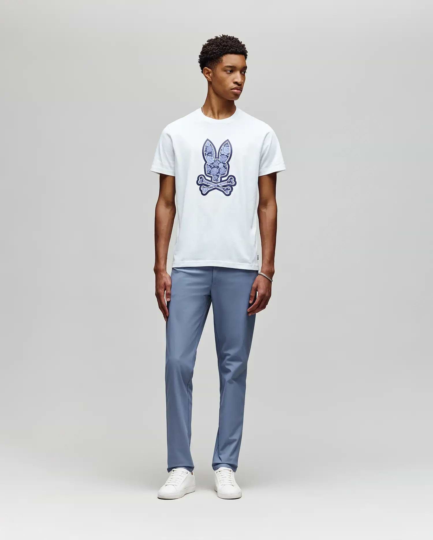 A young man stands against a light gray background, wearing the Psycho Bunny MENS ROWEN BUNNY GRAPHIC TEE (MT0200101) in white with a blue graphic, paired with light blue pants and white sneakers, looking slightly left with a neutral expression. (5)