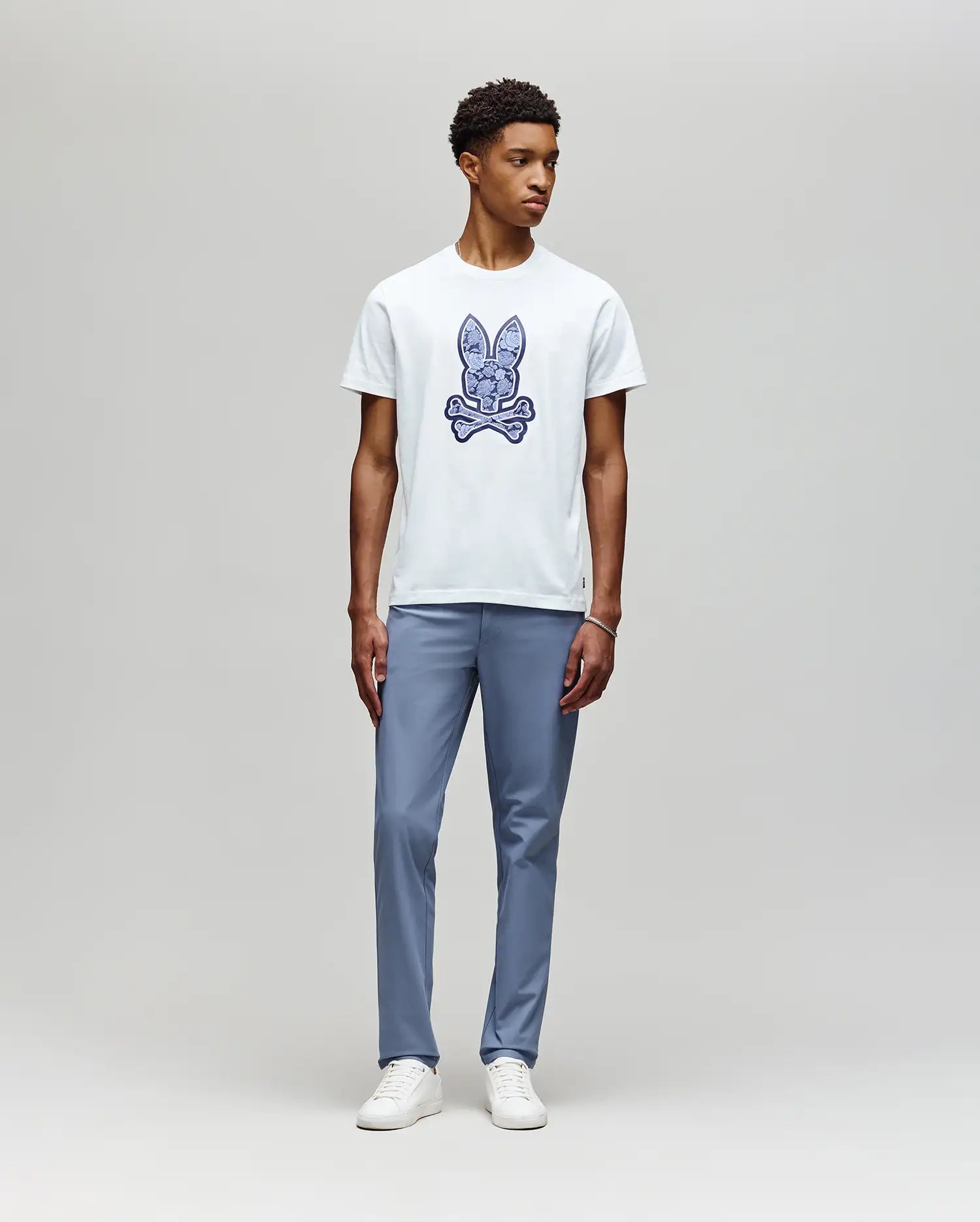 A young man stands against a light gray background, wearing the Psycho Bunny MENS ROWEN BUNNY GRAPHIC TEE (MT0200101) in white with a blue graphic, paired with light blue pants and white sneakers, looking slightly left with a neutral expression. (5)