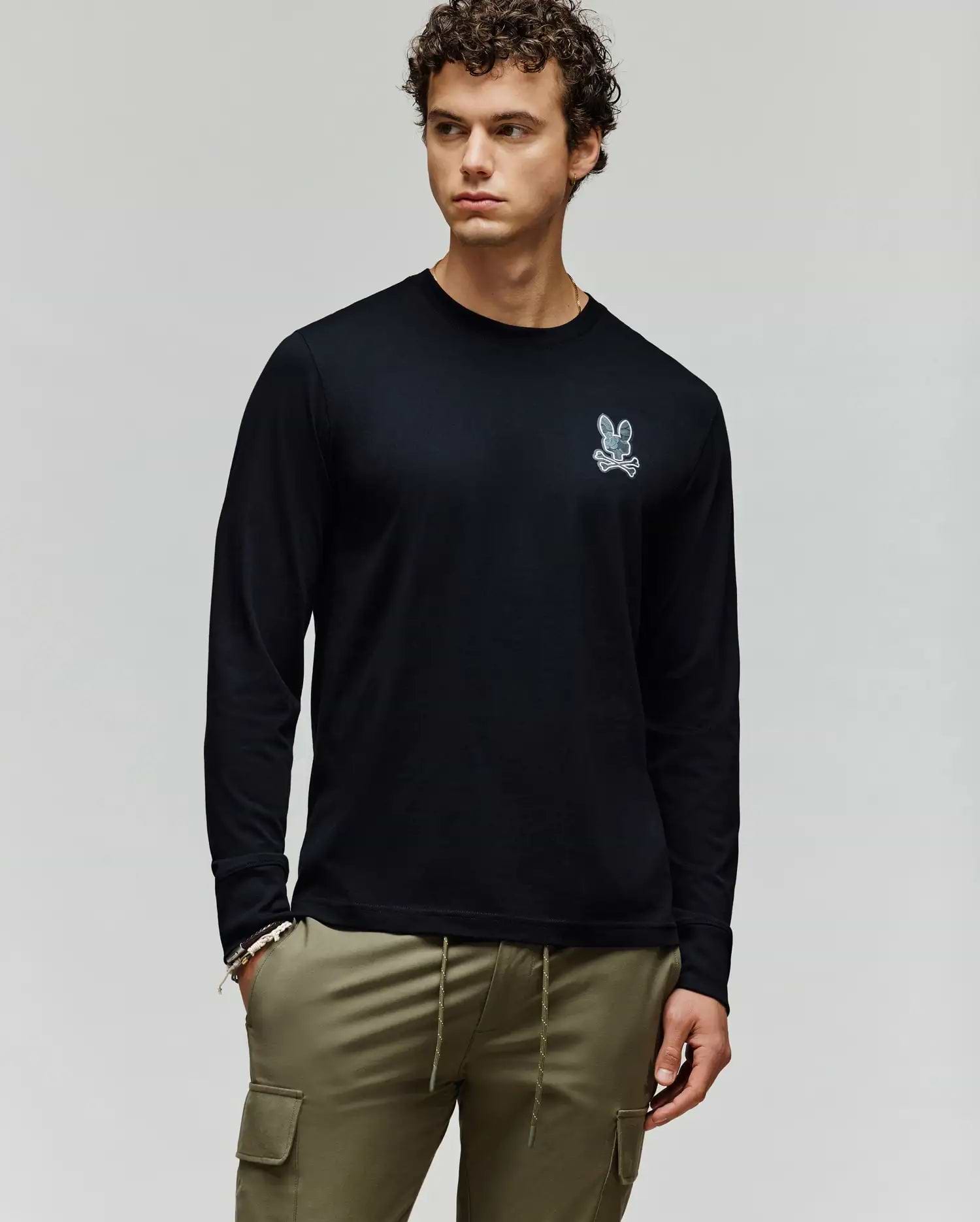 A man with curly hair wears the Psycho Bunny MENS ROWEN GRAPHIC TEE (MT0200103), a black long sleeve featuring a small white bunny logo on the chest, paired with olive green cargo pants against a plain light gray background. (1)