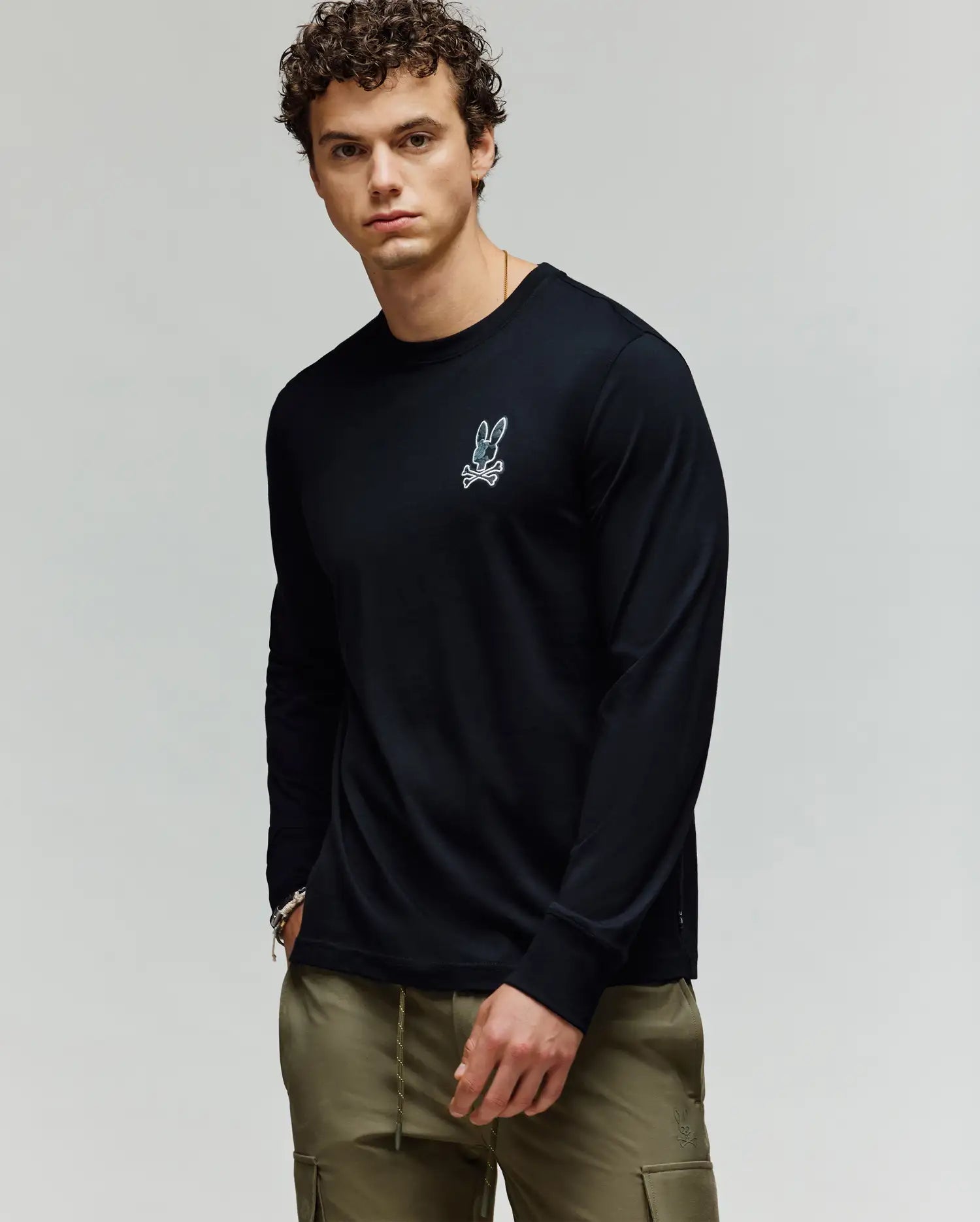 A man with curly hair wears the Psycho Bunny MENS ROWEN GRAPHIC TEE - MT0200103, a black long-sleeve shirt with a small white bunny logo on the chest, paired with olive green pants, standing against a plain light background. (2)