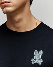A person wears the Psycho Bunny MENS ROWEN GRAPHIC TEE (MT0200103), a plain black Pima cotton shirt featuring a gray bunny and crossbones patch on the chest, paired with a thin gold chain necklace. Only the lower half of the face and neck are visible. (3)