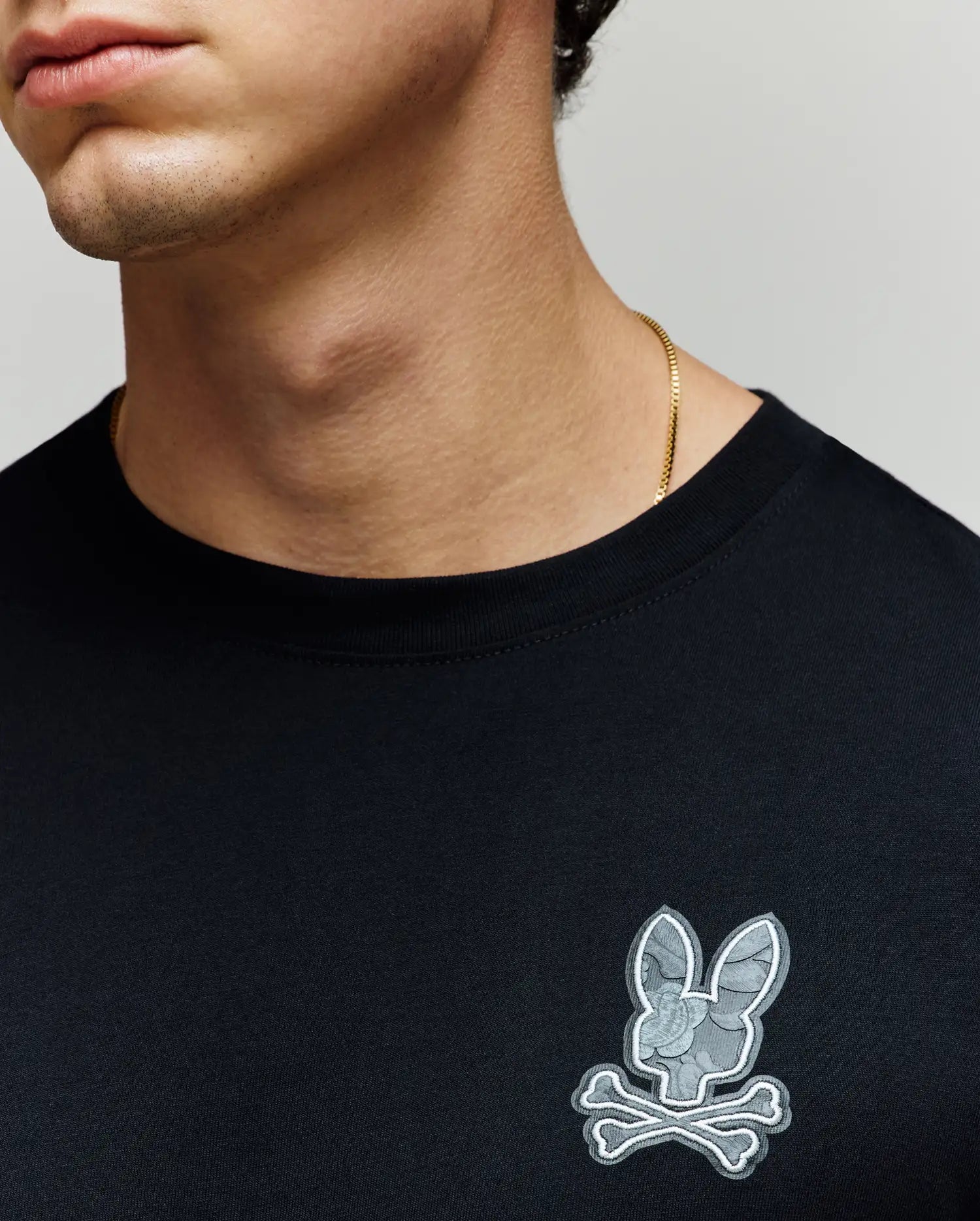A person wears the Psycho Bunny MENS ROWEN GRAPHIC TEE (MT0200103), a plain black Pima cotton shirt featuring a gray bunny and crossbones patch on the chest, paired with a thin gold chain necklace. Only the lower half of the face and neck are visible. (3)