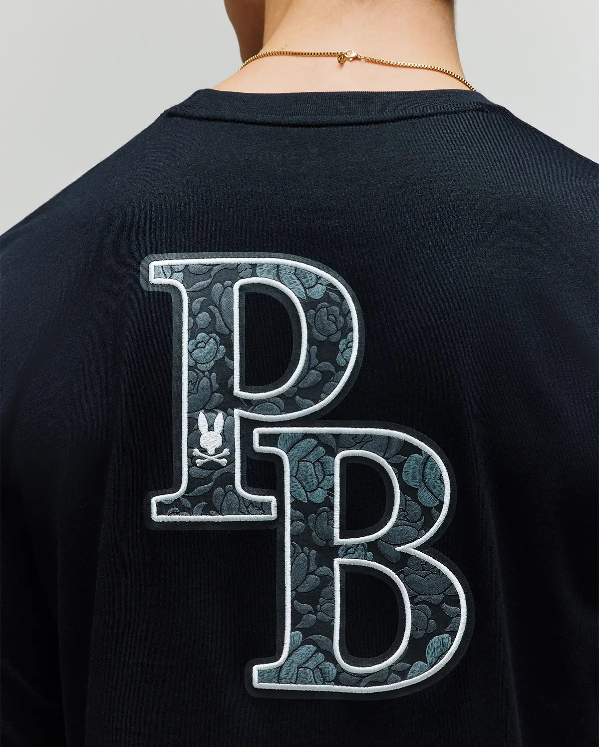A person wearing the Psycho Bunny MENS ROWEN GRAPHIC TEE (MT0200103), a black tee with large overlapping "P" and "B" rose-patterned graphics and a small bunny logo on the back. (4)