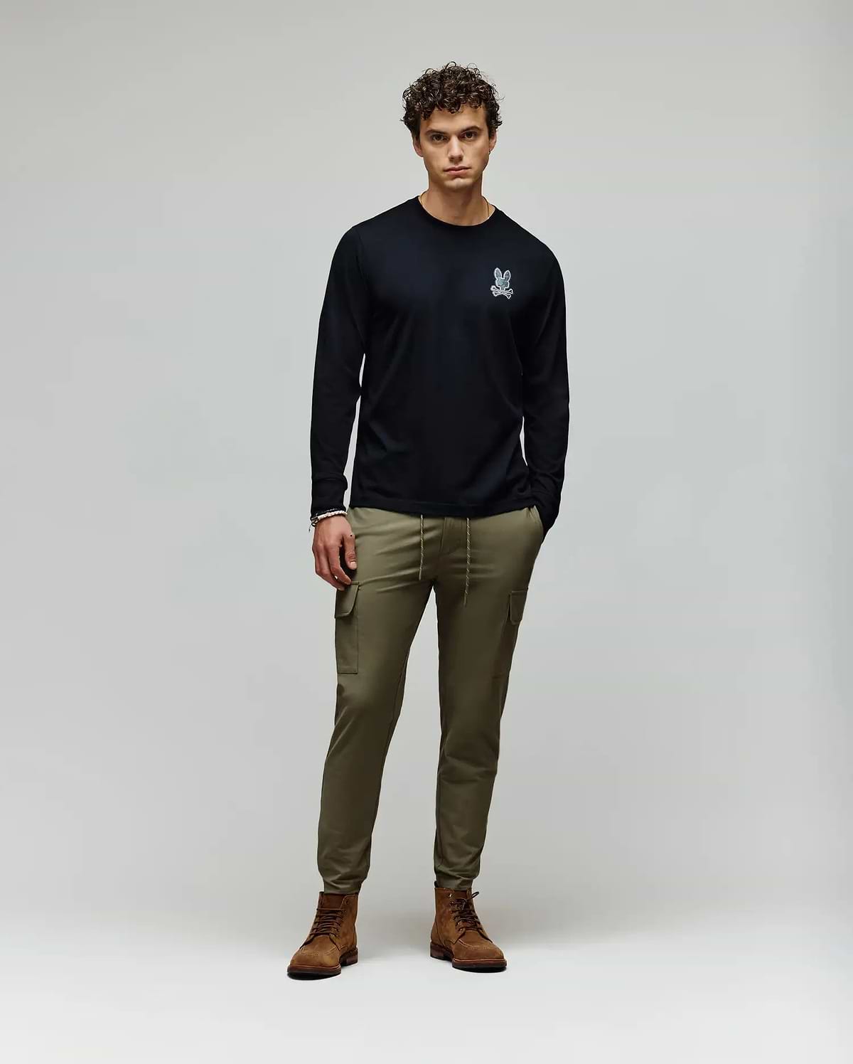 A man stands against a plain background wearing a black Psycho Bunny MENS ROWEN GRAPHIC TEE - MT0200103 with a small white chest graphic, olive green cargo pants, and brown boots, his hands in his pockets. (5)