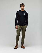 A man stands against a plain background wearing a black Psycho Bunny MENS ROWEN GRAPHIC TEE - MT0200103 with a small white chest graphic, olive green cargo pants, and brown boots, his hands in his pockets. (5)