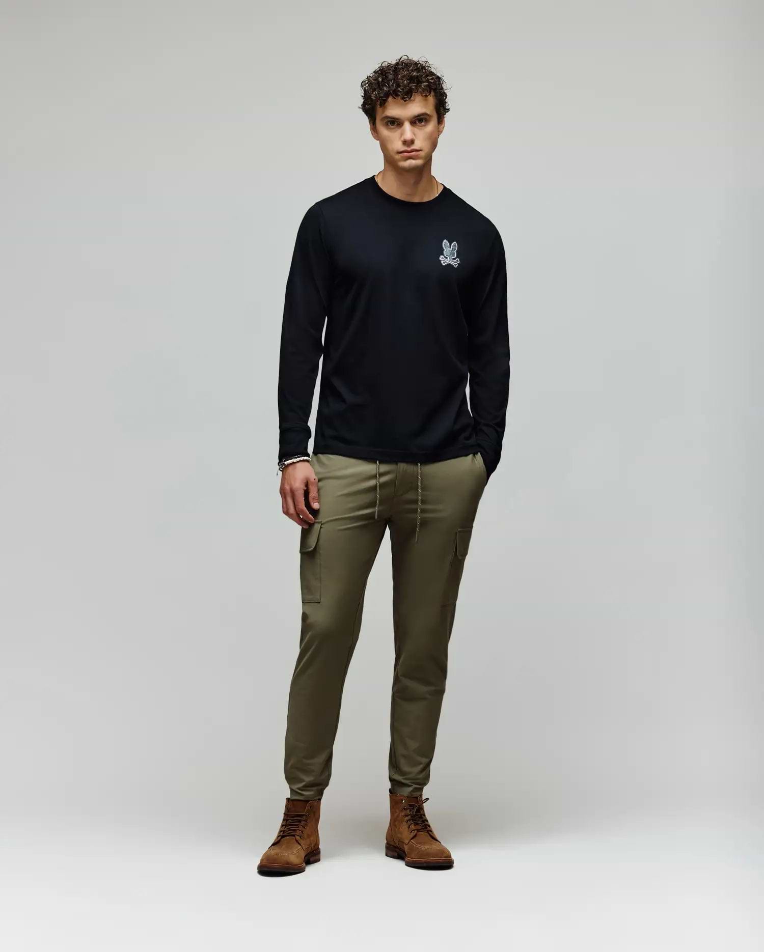 A man stands against a plain background wearing a black Psycho Bunny MENS ROWEN GRAPHIC TEE - MT0200103 with a small white chest graphic, olive green cargo pants, and brown boots, his hands in his pockets. (5)