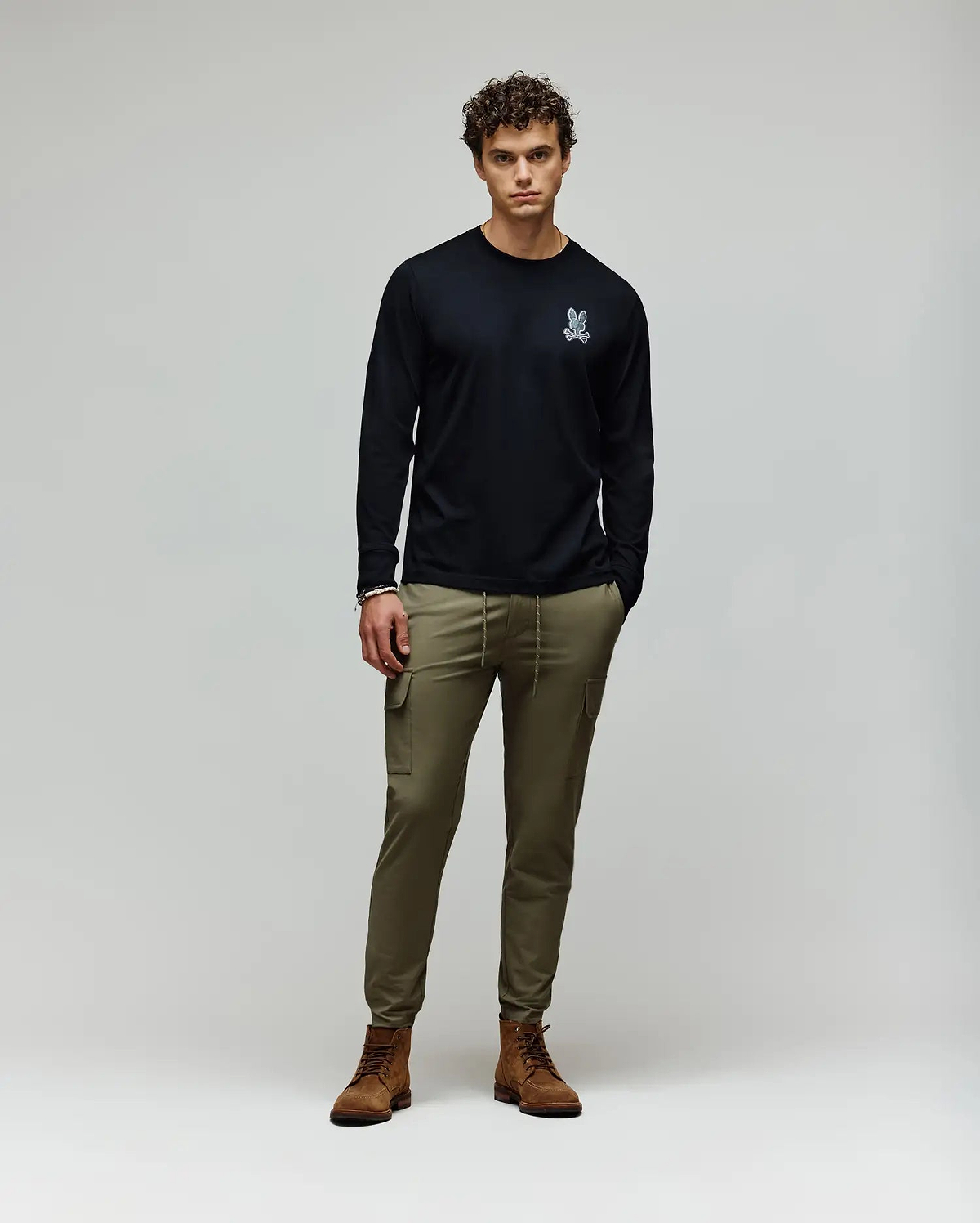 A man stands against a plain background wearing a black Psycho Bunny MENS ROWEN GRAPHIC TEE - MT0200103 with a small white chest graphic, olive green cargo pants, and brown boots, his hands in his pockets. (5)