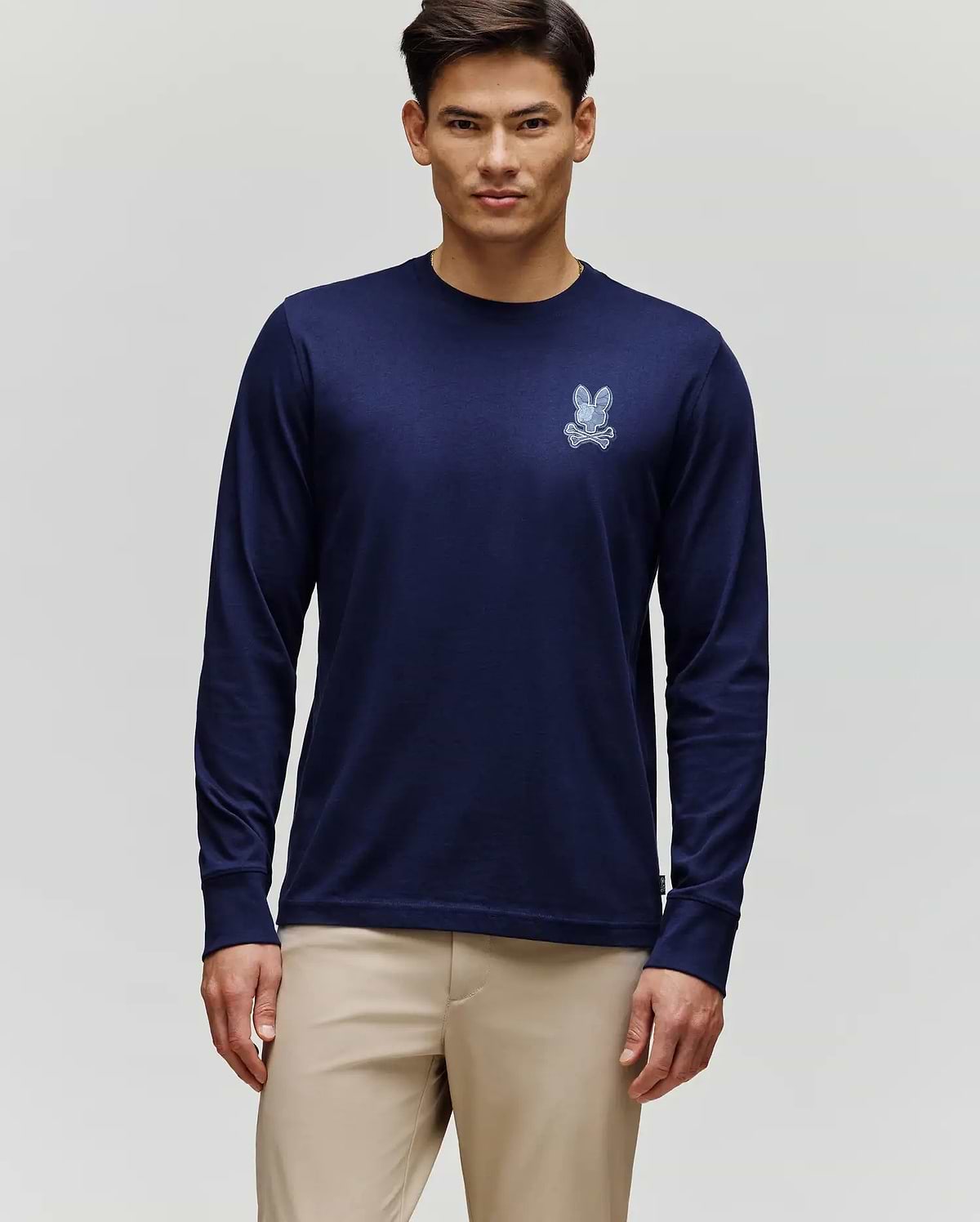 A man stands against a light gray background wearing Psycho Bunny’s MENS ROWEN GRAPHIC TEE - MT0200103 in navy blue with a small white graphic on the left chest, paired with beige pants.