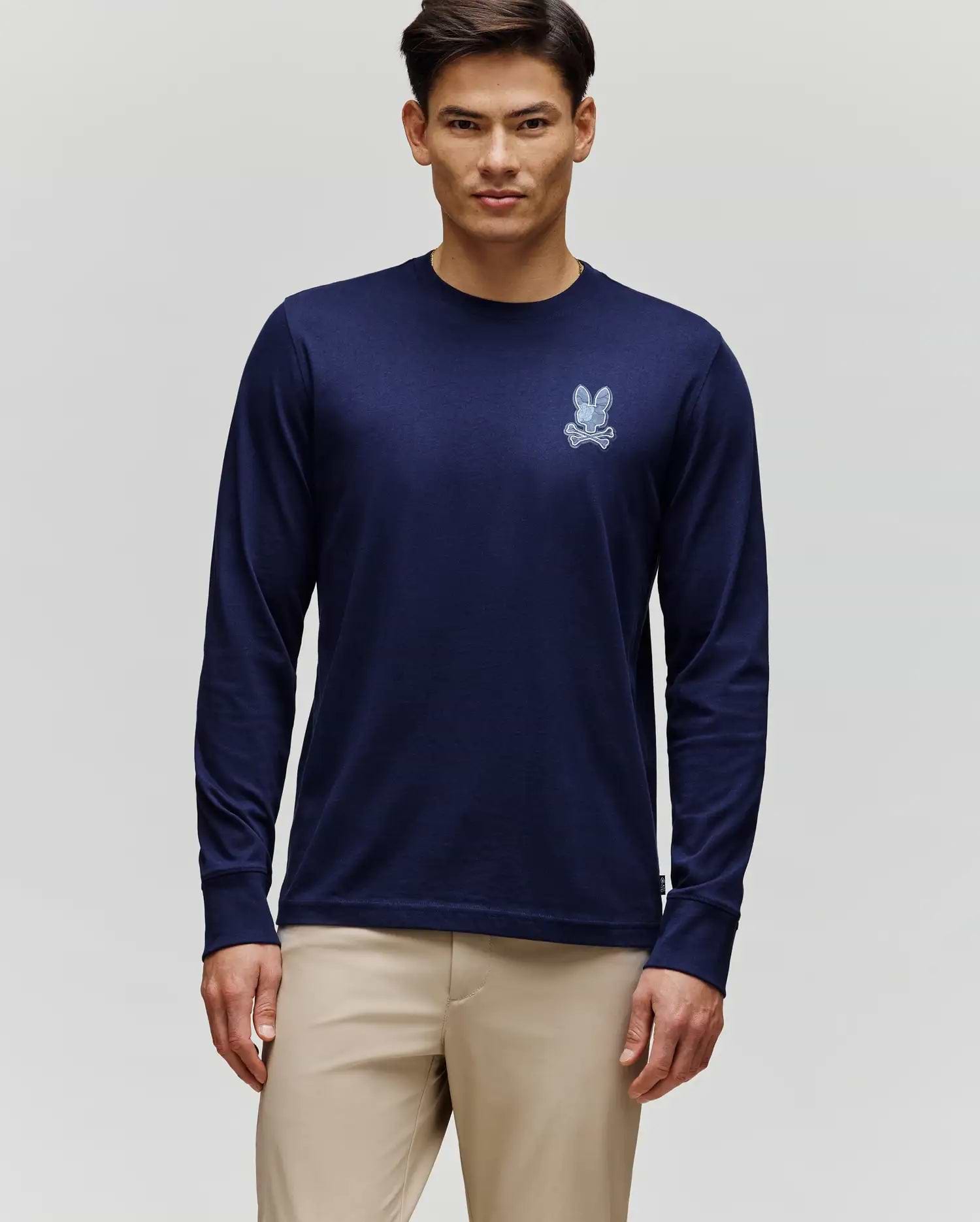 A man stands against a light gray background wearing Psycho Bunny’s MENS ROWEN GRAPHIC TEE - MT0200103 in navy blue with a small white graphic on the left chest, paired with beige pants.