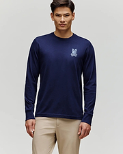 A man stands against a light gray background wearing Psycho Bunny’s MENS ROWEN GRAPHIC TEE - MT0200103 in navy blue with a small white graphic on the left chest, paired with beige pants.