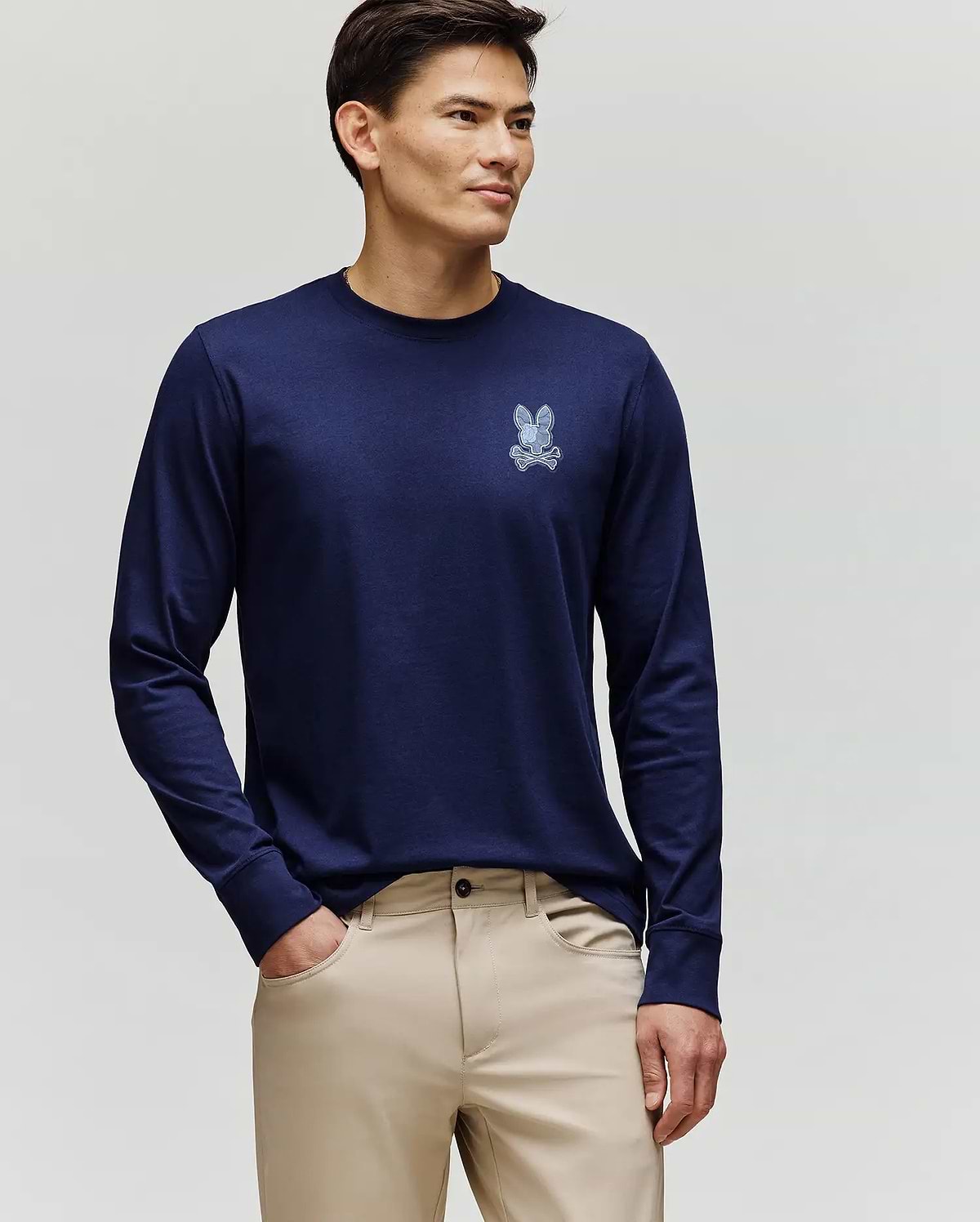 A man faces slightly left, wearing the Psycho Bunny MENS ROWEN GRAPHIC TEE - MT0200103 in navy blue with a small chest bunny graphic and beige pants, against a plain light background.