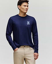 A man faces slightly left, wearing the Psycho Bunny MENS ROWEN GRAPHIC TEE - MT0200103 in navy blue with a small chest bunny graphic and beige pants, against a plain light background.