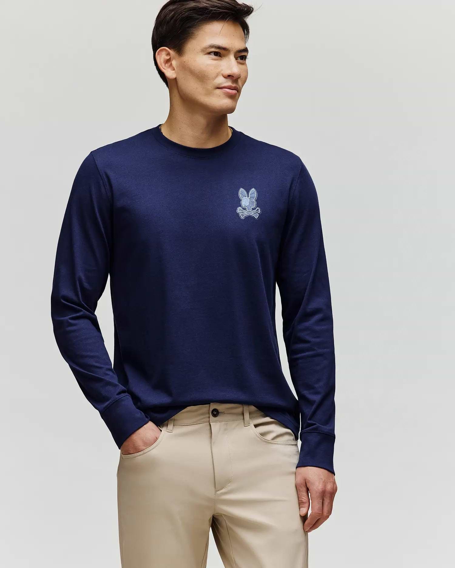 A man faces slightly left, wearing the Psycho Bunny MENS ROWEN GRAPHIC TEE - MT0200103 in navy blue with a small chest bunny graphic and beige pants, against a plain light background.
