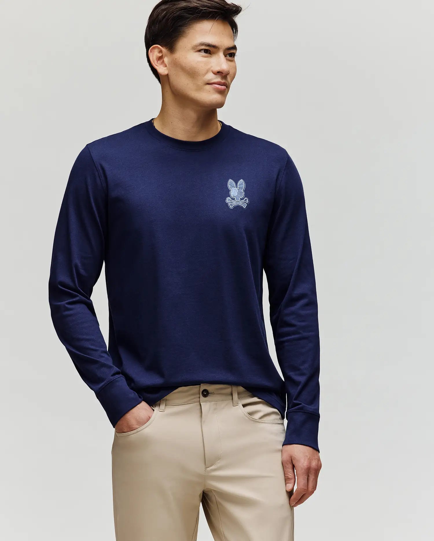 A man faces slightly left, wearing the Psycho Bunny MENS ROWEN GRAPHIC TEE - MT0200103 in navy blue with a small chest bunny graphic and beige pants, against a plain light background.