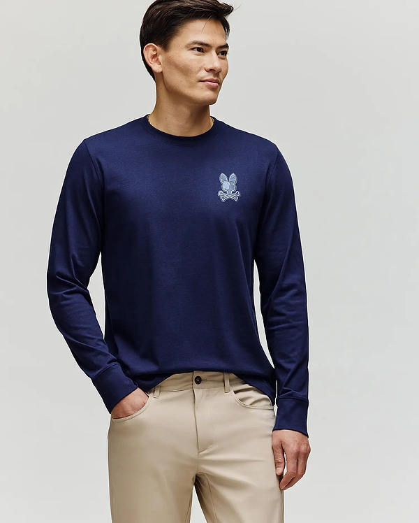 A man faces slightly left, wearing the Psycho Bunny MENS ROWEN GRAPHIC TEE - MT0200103 in navy blue with a small chest bunny graphic and beige pants, against a plain light background.