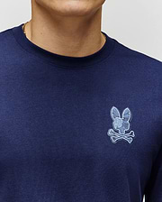 Close-up of a person wearing the Psycho Bunny MENS ROWEN GRAPHIC TEE (MT0200103) in navy blue Pima cotton, featuring a light blue bunny skull and crossbones logo on the left chest; only lower face and upper torso are visible.