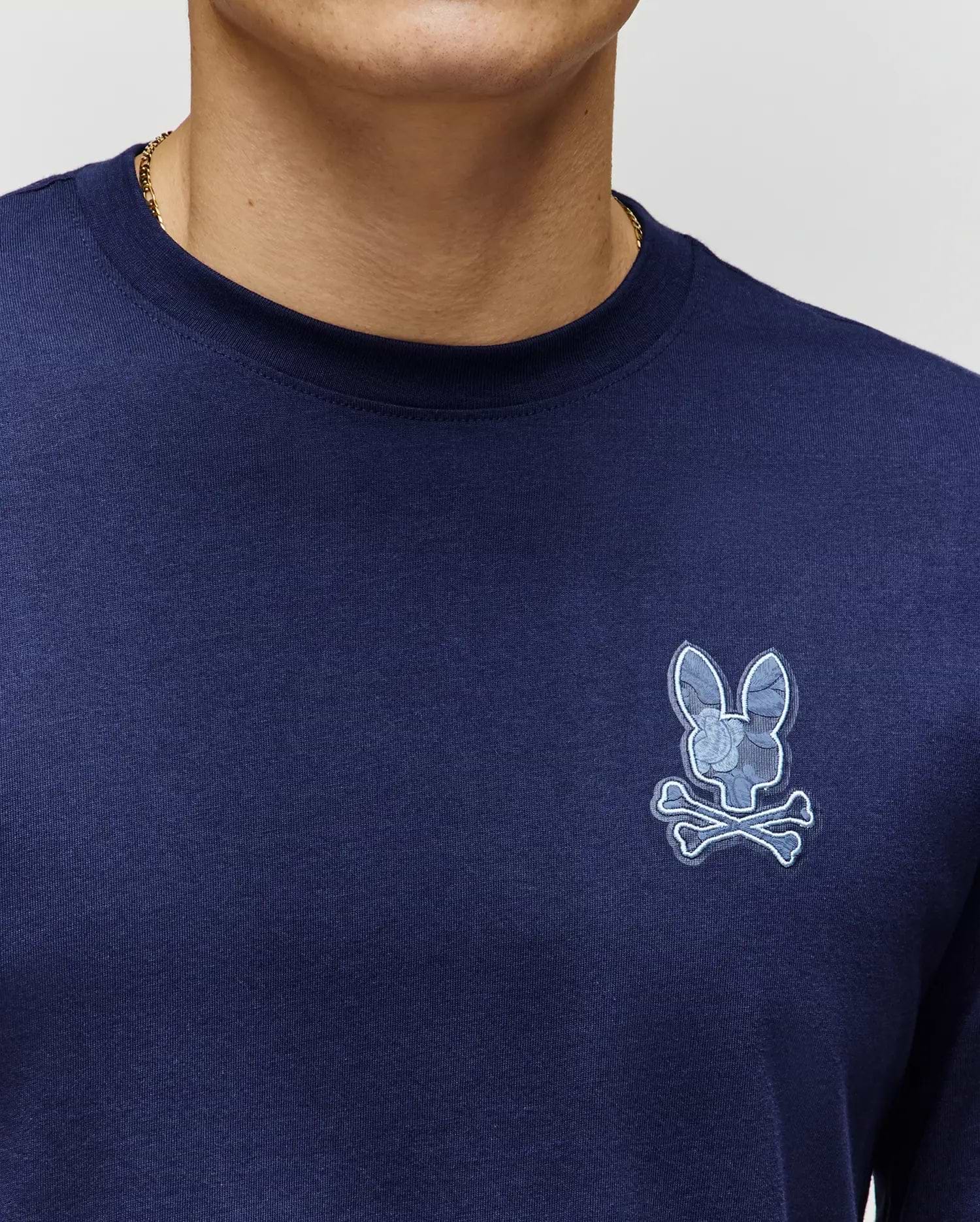 Close-up of a person wearing the Psycho Bunny MENS ROWEN GRAPHIC TEE (MT0200103) in navy blue Pima cotton, featuring a light blue bunny skull and crossbones logo on the left chest; only lower face and upper torso are visible.