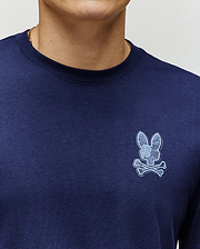 Close-up of a person wearing the Psycho Bunny MENS ROWEN GRAPHIC TEE (MT0200103) in navy blue Pima cotton, featuring a light blue bunny skull and crossbones logo on the left chest; only lower face and upper torso are visible.