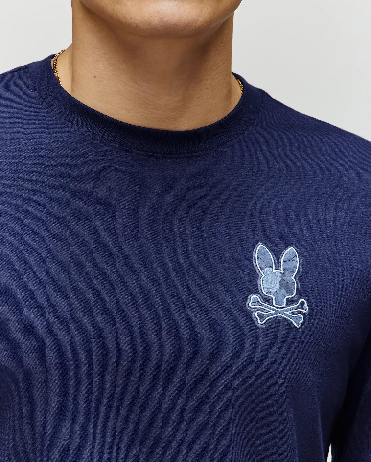 Close-up of a person wearing the Psycho Bunny MENS ROWEN GRAPHIC TEE (MT0200103) in navy blue Pima cotton, featuring a light blue bunny skull and crossbones logo on the left chest; only lower face and upper torso are visible.