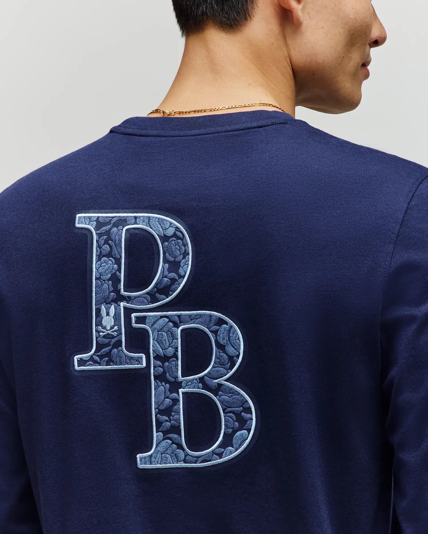 A person wears the Psycho Bunny MENS ROWEN GRAPHIC TEE (MT0200103), a navy long sleeve with large, patterned "PB" floral letters outlined in light blue on the back. Only part of their face is visible as they're shown from behind.