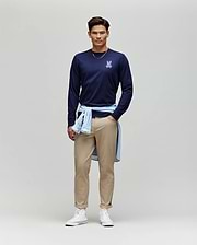 A man stands against a plain background, wearing the Psycho Bunny MENS ROWEN GRAPHIC TEE - MT0200103 in navy, tan pants, white high-top sneakers, and a light blue shirt tied around his waist.
