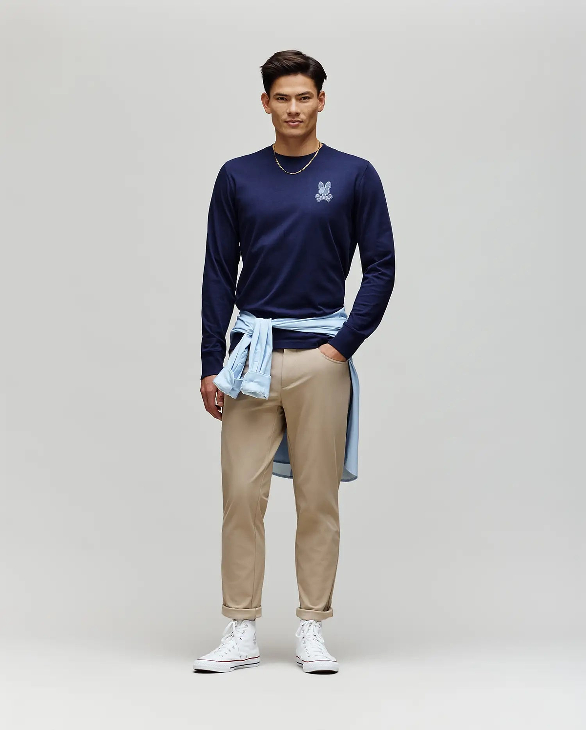 A man stands against a plain background, wearing the Psycho Bunny MENS ROWEN GRAPHIC TEE - MT0200103 in navy, tan pants, white high-top sneakers, and a light blue shirt tied around his waist.