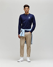 A man stands against a plain background, wearing the Psycho Bunny MENS ROWEN GRAPHIC TEE - MT0200103 in navy, tan pants, white high-top sneakers, and a light blue shirt tied around his waist.