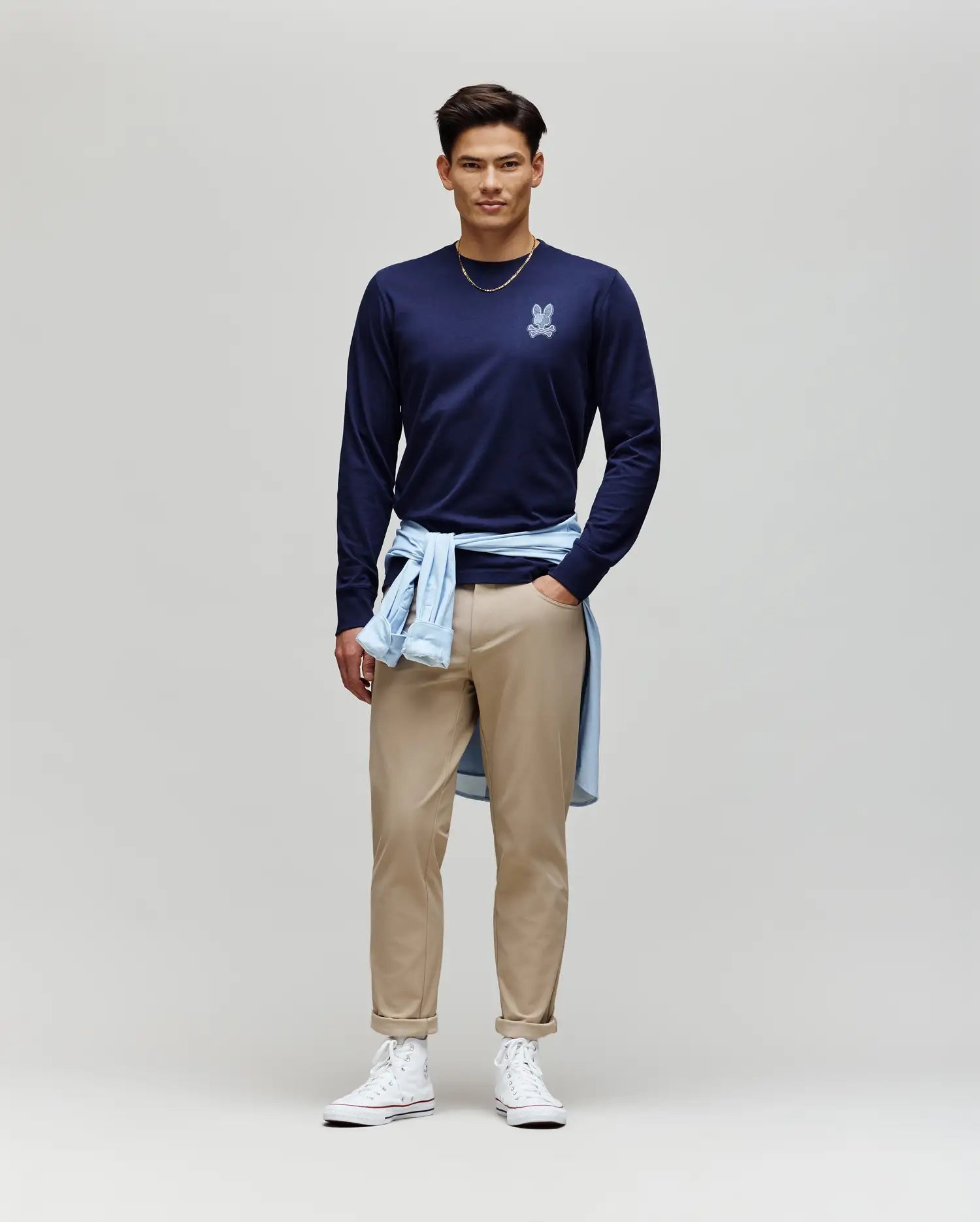 A man stands against a plain background, wearing the Psycho Bunny MENS ROWEN GRAPHIC TEE - MT0200103 in navy, tan pants, white high-top sneakers, and a light blue shirt tied around his waist.