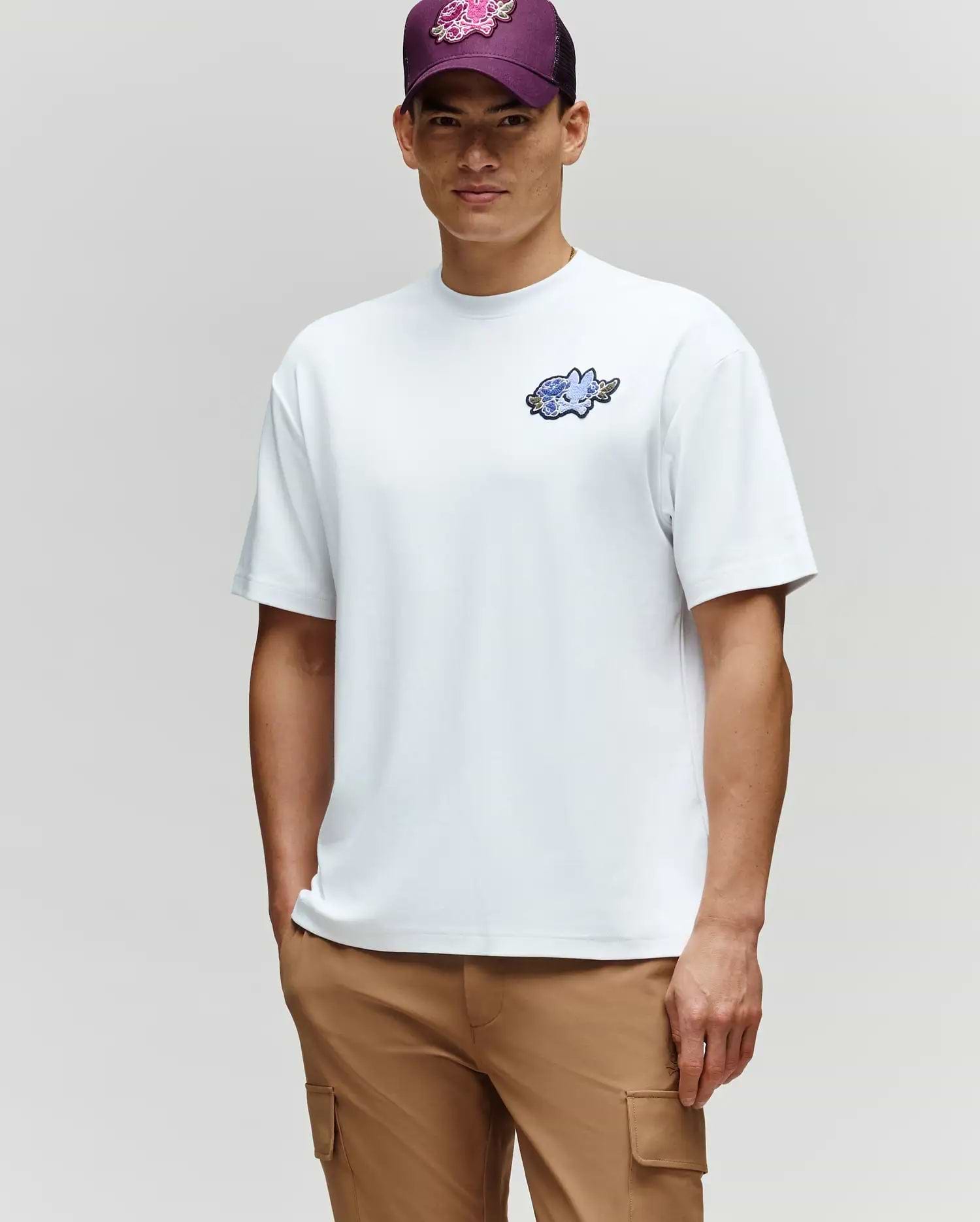 A man exudes laidback style in the Psycho Bunny MENS AUGUSTE OVERSIZED TEE (MT0200104), tan cargo pants, and a purple floral cap as he stands against a light gray background.