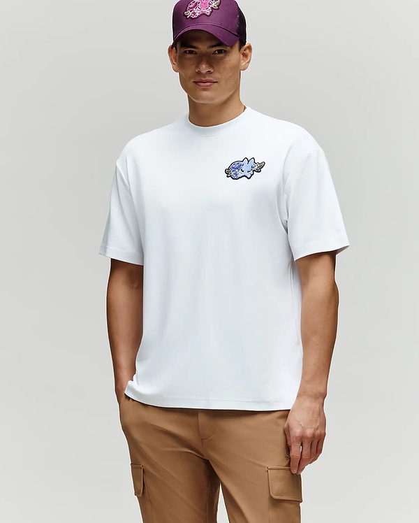 A man exudes laidback style in the Psycho Bunny MENS AUGUSTE OVERSIZED TEE (MT0200104), tan cargo pants, and a purple floral cap as he stands against a light gray background.
