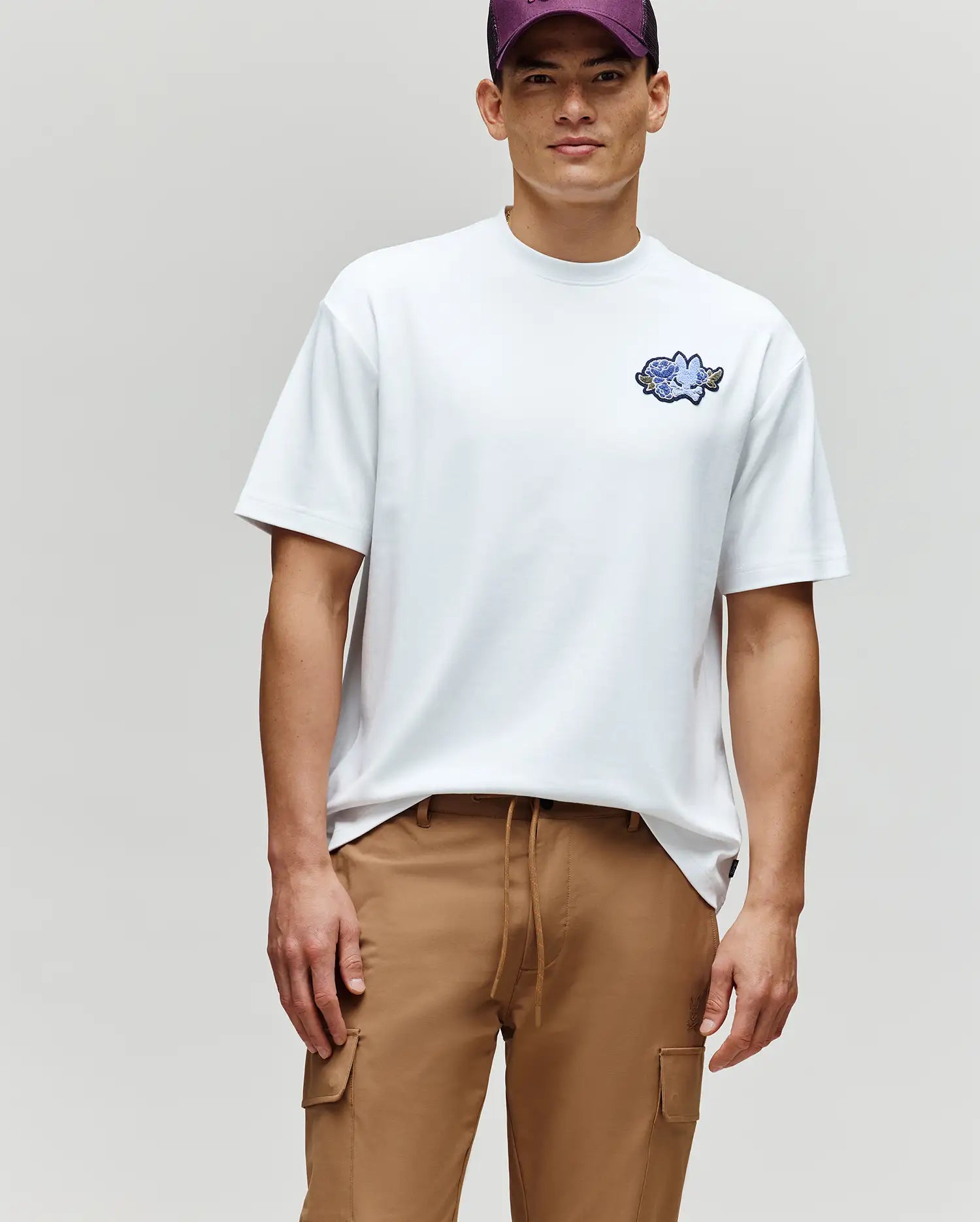 A man in the Psycho Bunny MENS AUGUSTE OVERSIZED TEE (MT0200104), featuring a small blue cartoon fish graphic, tan cargo pants, and a purple cap stands against a plain background, embodying an easygoing, laidback style.