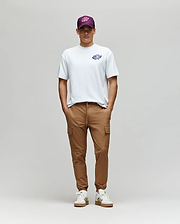 A man stands against a plain background in the Psycho Bunny MENS AUGUSTE OVERSIZED TEE (MT0200104) with an embroidered Bunny logo, paired with tan cargo joggers, white sneakers, and a purple cap for a laidback look.