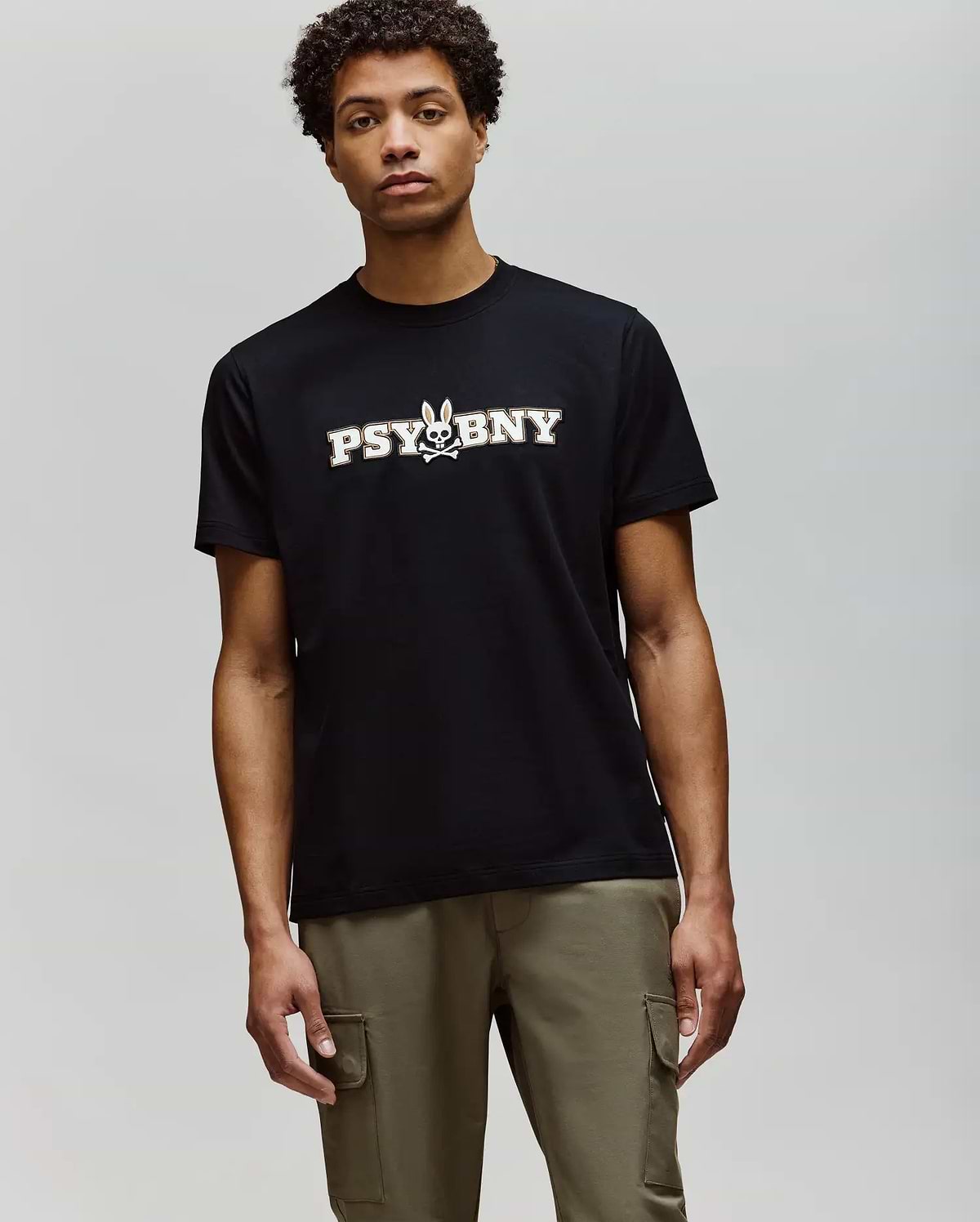 A man wears the Psycho Bunny MENS WADE GRAPHIC TEE (MT0200105), a regular fit black tee with "PSYBNY" and a small bunny graphic, plus olive green cargo pants. The 100% cotton shirt offers comfort against a plain background. (2)