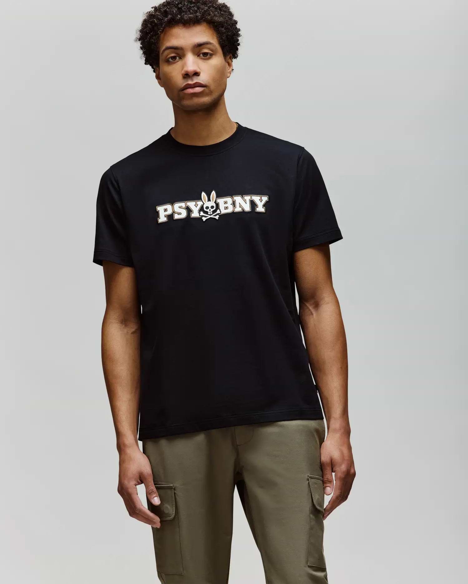 A man wears the Psycho Bunny MENS WADE GRAPHIC TEE (MT0200105), a regular fit black tee with "PSYBNY" and a small bunny graphic, plus olive green cargo pants. The 100% cotton shirt offers comfort against a plain background. (2)