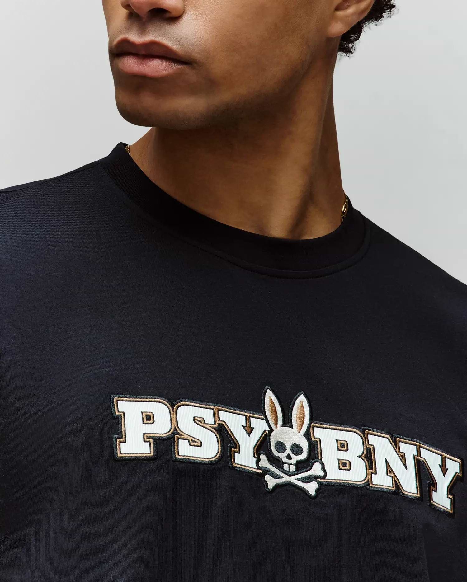 Wearing Psycho Bunny’s MENS WADE GRAPHIC TEE (MT0200105), a black 100% cotton regular fit shirt with “PSYBNY” text and a skull with bunny ears and crossbones logo; only the lower face and upper torso are visible. (3)